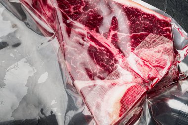 T-bone ya da Floransa kuru çiğ mermer biftek vakum ambalaj seti, siyah taş arka planda, üst görünüm düz yatıyordu