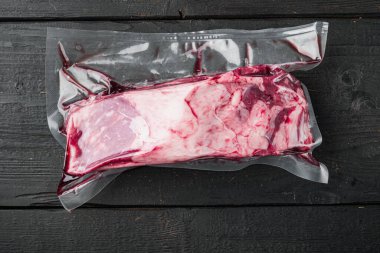 Süpürge siyah plastik paket ve taze biftek. Etiketsiz paketlenmiş çiğ et, siyah ahşap masa arkaplanı, üst görünüm düz yatıyordu