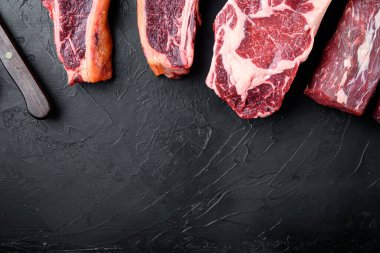 Baharat ve çatal bıçak seti, tomahawk, t kemik, sopa biftek, pirzola ve fileto kesikleri ile ızgara için çiğ et bifteği çeşitleri, siyah taş arka planda, üst düz görünüm yatağı, metin için kopya alanı