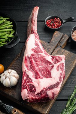 Çiğ taze tomahawk kara angus dana pirzolası seti, ızgara malzemeli, siyah ahşap masa arka planında