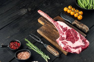Piece of raw rib eye tomahawk or cowboy steak, raw fresh marbled prime beef set, with grill ingredients, on black wooden table background, with copy space for text clipart