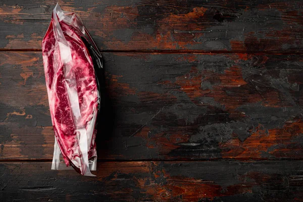 Vacuum black plastic pack with fresh beef steak. Raw meat packed without label set, on old dark  wooden table background, top view flat lay, with copy space for text