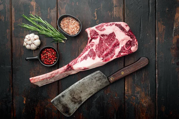 Raw uncooked black angus beef tomahawk steak on bone set, and old butcher cleaver knife, with seasoning and herbs, on old dark  wooden table background, top view flat lay, with copy space for text