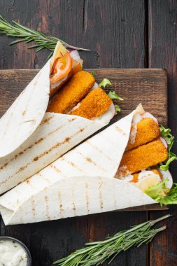 Balık kroketli tortilla rulosu, peynir ve sebze seti, ahşap kesme tahtasının üzerinde, eski siyah ahşap masa zemininde, üst manzara düz