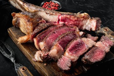 Kemik dilimlenmiş ızgara biftek, orta pişmiş balta kesim seti, ahşap servis tahtası, siyah taş arka plan.