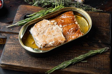 Tütsülenmiş somon filetosu konserve balık seti, ahşap kesme tahtası, eski, siyah ahşap masa arka planında bitkiler ve malzemelerle birlikte.