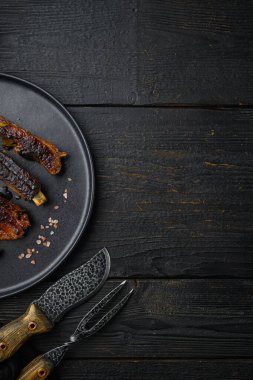 Izgara domuz pirzolası, tabakta, barbekü bıçağı ve et çatalı, siyah ahşap masa zemininde, üst manzara düz, metin için kopyalama alanı var.