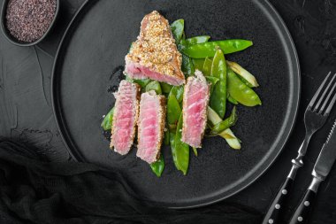 Taze soğan ve bezelyeli ızgara kırmızı nadir ton balığı bifteği. Tabakta, siyah taş zemin üzerinde. Üst manzara düz.