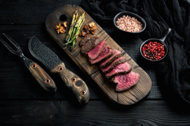 Izgara fileminyon biftek seti, soğan ve kuşkonmaz, ahşap servis tahtasında, et bıçağı ve çatalla, siyah ahşap masa arkasında.