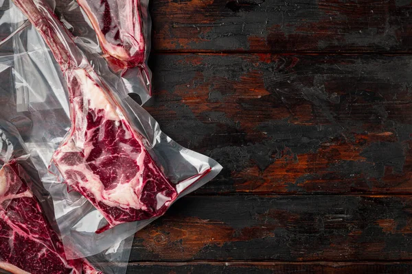 Dry aged steak in a vacuum. Meat products in plastic pack set, tomahawk, t bone and club steak cuts, on old dark  wooden table background, top view flat lay, with copy space for text