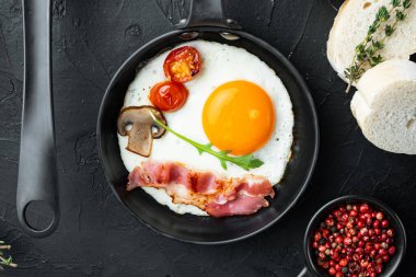 Kızarmış yumurta, kiraz domatesli ve kahvaltıda ekmek, dökme demir tavada, siyah arka planda, üst manzara düz.