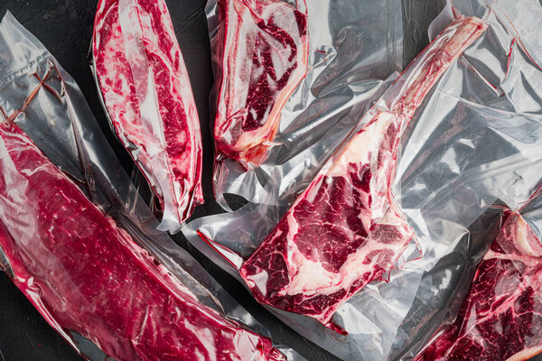 Raw marbled beef steak in vacuum packaging set, tomahawk, t bone, club steak, rib eye and tenderloin cuts, on black stone background, top view flat lay