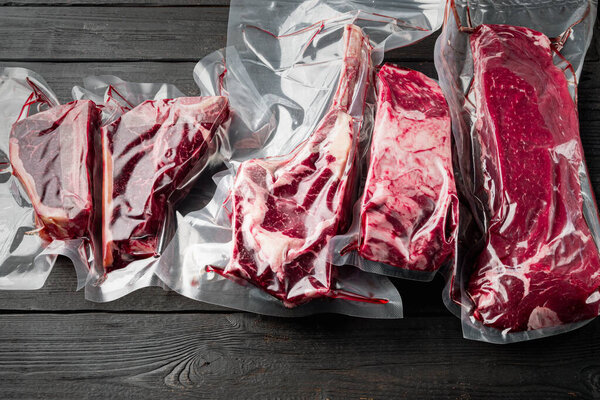 Raw marbled beef steak in vacuum packaging set, tomahawk, t bone, club steak, rib eye and tenderloin cuts, on black wooden table background
