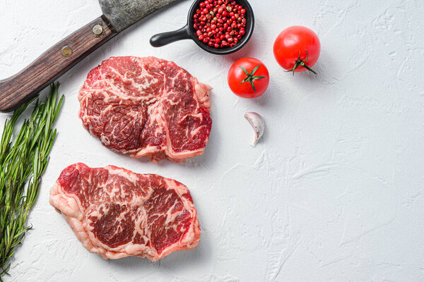 Organic top blade steak, raw beef meat with seasonings, rosemary and butcher cleaver. White textured background. Top view space for text