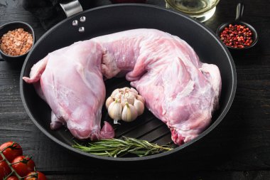 Baharatlı ve malzemeli çiğ tavşan eti, dökme demir tavada, siyah ahşap masa arkasında.