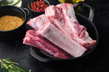 Taze çiğ domuz kaburgaları ve malzemeler, kızarmış demir tavada, siyah taş arka planda.