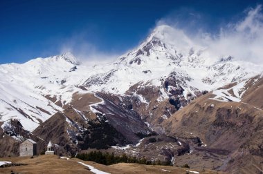 Arka planda karla kaplı Kazbegi dağlı küçük ortodoks kilisesi, açık mavi gökyüzü.