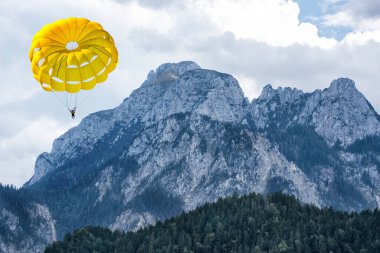 Yüksek Alpler dağlarının arka planında bir paraşütle süzülüyordu. Mavi bulutlu gökyüzünün altında yeşil bir orman vardı..