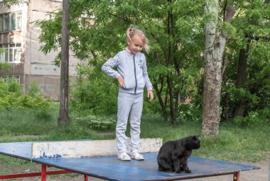 Küçük bir kız, beş yaşında Avrupalı, parkta bir tenis masasında duruyor, ve yanında siyah bir sokak kedisi oturuyor.