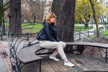 50 yaşında, orta yaşlı, spor ceketli ve eşofmanlı bir kadın. Bir bankta oturuyor, bahar manzarası var.