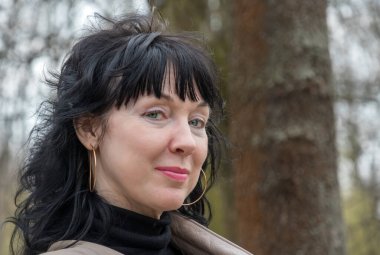 Bayan portresi, orta yaşlı güzel bir kadın, iri gözlü, uzun siyah saçlı ve neşeli bakışlı, parkta duruyor.