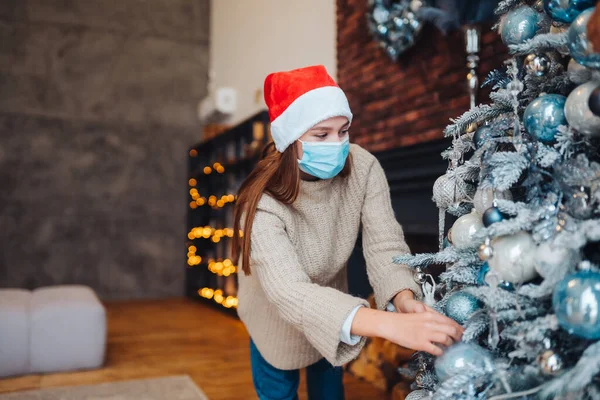 Seorang wanita muda menghias pohon Natal dengan masker medis. — Stok Foto