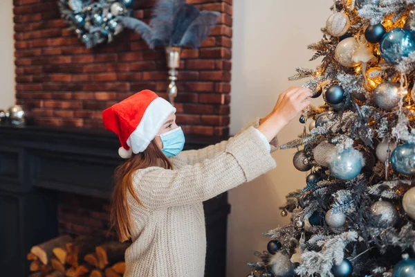 Seorang wanita muda menghias pohon Natal dengan masker medis. — Stok Foto