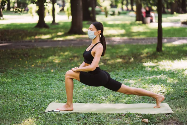 Giovane donna atletica in una maschera protettiva medica, facendo yoga nel parco — Foto Stock