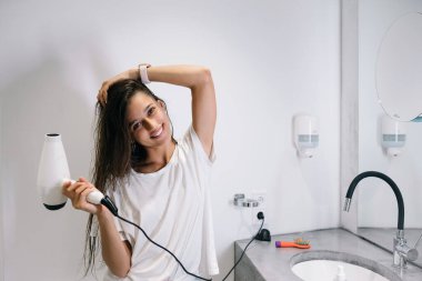 Genç ve güzel bir kadın banyoda elinde saç kurutma makinesi tutuyor.