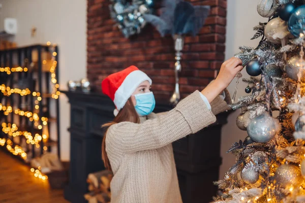 Seorang wanita muda menghias pohon Natal dengan masker medis. — Stok Foto