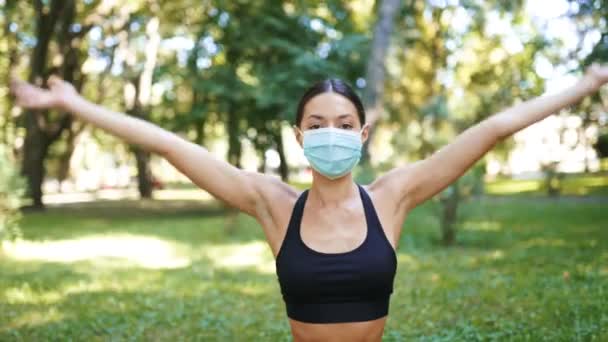 Sportowa młoda kobieta w medycznej masce ochronnej, uprawiająca jogę w parku — Wideo stockowe