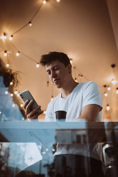 Młody, przystojny mężczyzna używa smartfona w kawiarni. Zdjęcie za szkłem — Zdjęcie stockowe