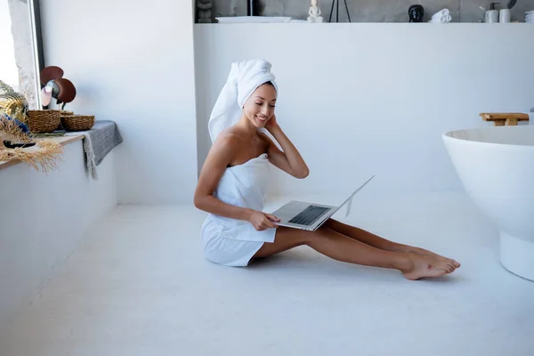 Donna in asciugamano bianco si siede in bagno e utilizzare un computer portatile. — Foto Stock