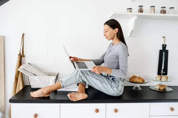 Bild der netten brünetten Frau, die in der Küche sitzt und Laptop benutzt — Stockfoto