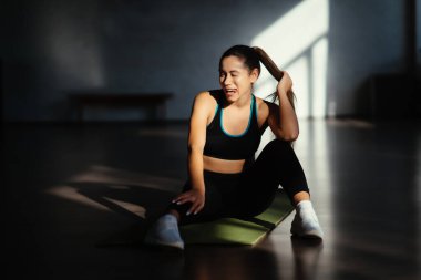 Formda bir kadın antrenmandan sonra dinleniyor, paspasın üzerinde oturuyor.