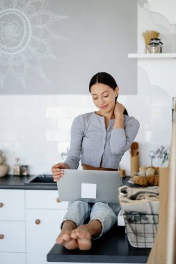 Şirin esmer bir kadının mutfakta oturup dizüstü bilgisayar kullanması.