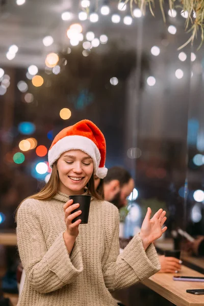 Dívka v klobouku Santa Claus sedí v kavárně a pije kávu — Stock fotografie