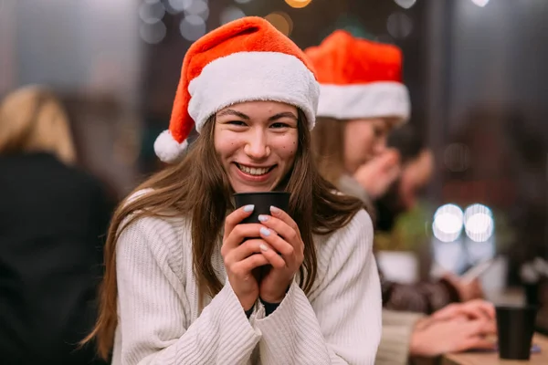 Dívka v klobouku Santa sedí v kavárně a pije kávu — Stock fotografie