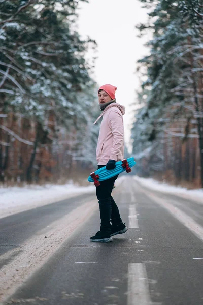Skateboarder de pé na estrada no meio da floresta, cercado por neve — Fotografia de Stock