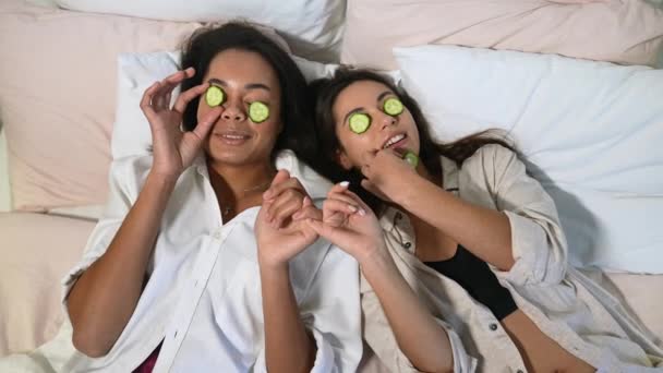 Otthoni fürdő. Két nő egy darab uborkát tart az arcán, az ágyon fekve.. — Stock videók