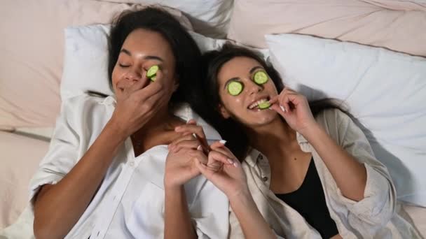 Thuis spa. Twee vrouwen met stukjes komkommer op hun gezicht op het bed. — Stockvideo