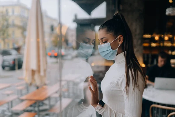 Молодая женщина в маске стоит перед окнами в кафе. — стоковое фото
