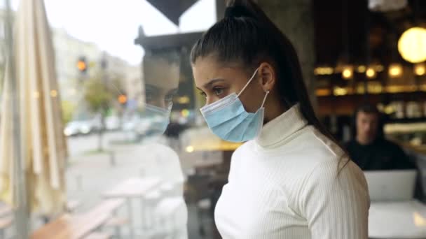 Mujer joven con mascarilla de pie frente a las ventanas en la cafetería. — Vídeos de Stock