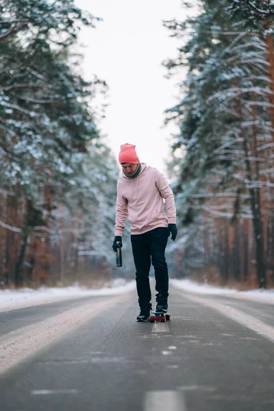 Skateboarder de pé na estrada no meio da floresta, cercado por neve — Fotografia de Stock