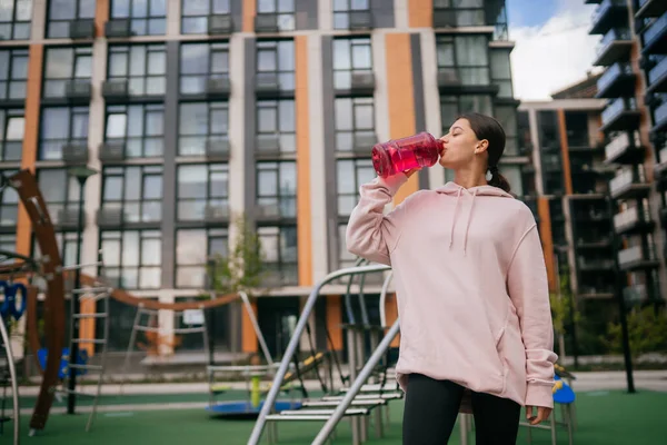 Una joven hermosa mujer está bebiendo agua en el patio de recreo — Foto de Stock