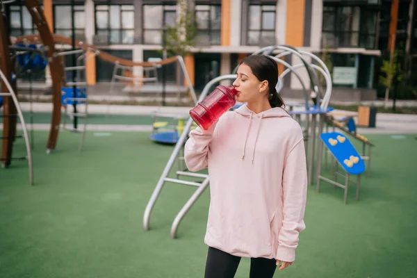 Una joven hermosa mujer está bebiendo agua en el patio de recreo —  Fotos de Stock