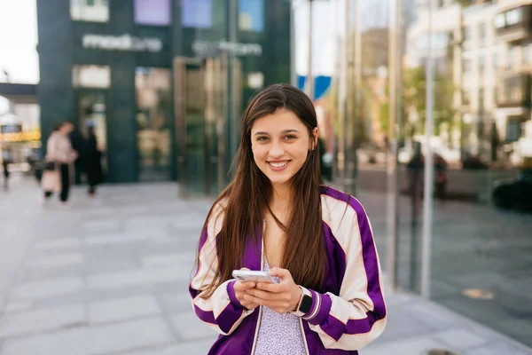 Sokaktaki mutlu kadın akıllı telefon kullanıyor ve kameraya bakıyor. — Stok fotoğraf