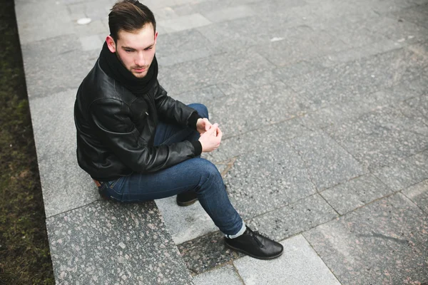 Um homem vestido de jeans e assentos de jaqueta preta em uma laje — Fotografia de Stock