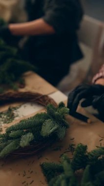 Toplum çalışma grubu masanın üzerinde çelenk yapıyor, katılımcılar eldiven takıyor ve asma köküne çam filizi dokumak için işbirliği yapıyorlar.