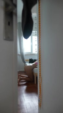 Yatak odasının pencerenin yanında diz çökmüş bir kadının kapı önü. Perde çerçevelenmiş, güneşli ahşap zemin ve küçük bir başucu taburesi.